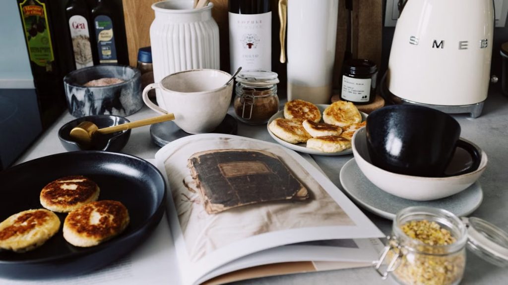 One portion of homemade cheese pancakes in frying pan and another on plate locating near opened stylish magazine among kitchen utensils and appliances in bright kitchen at home