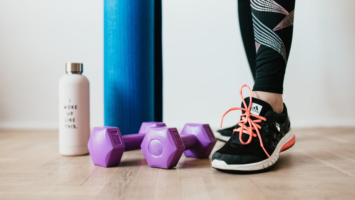 Crop sportswoman wearing sneakers and black leggings standing with sport mat on wooden floor near dumbbells and water bottle before exercising indoors